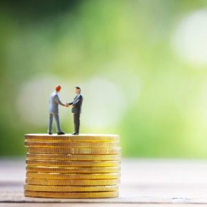 積み重ねた金貨の上に握手をしながら立つ2人の男性のイメージ